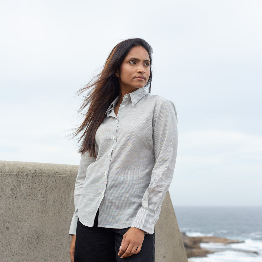 A woman wearing a light gray linen button-down shirt and black pants, standing outdoors near a concrete barrier with the ocean and cloudy sky in the background.